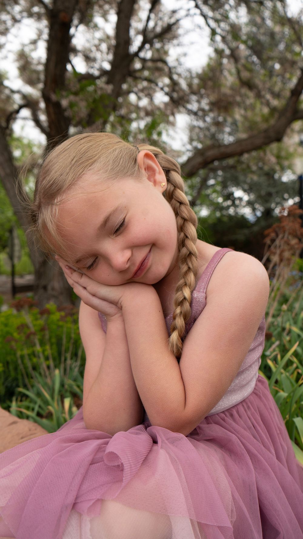 A young girl with a blonde braid wearing a pink tulle dress sits outdoors, smiling with hands clasped under her cheek.