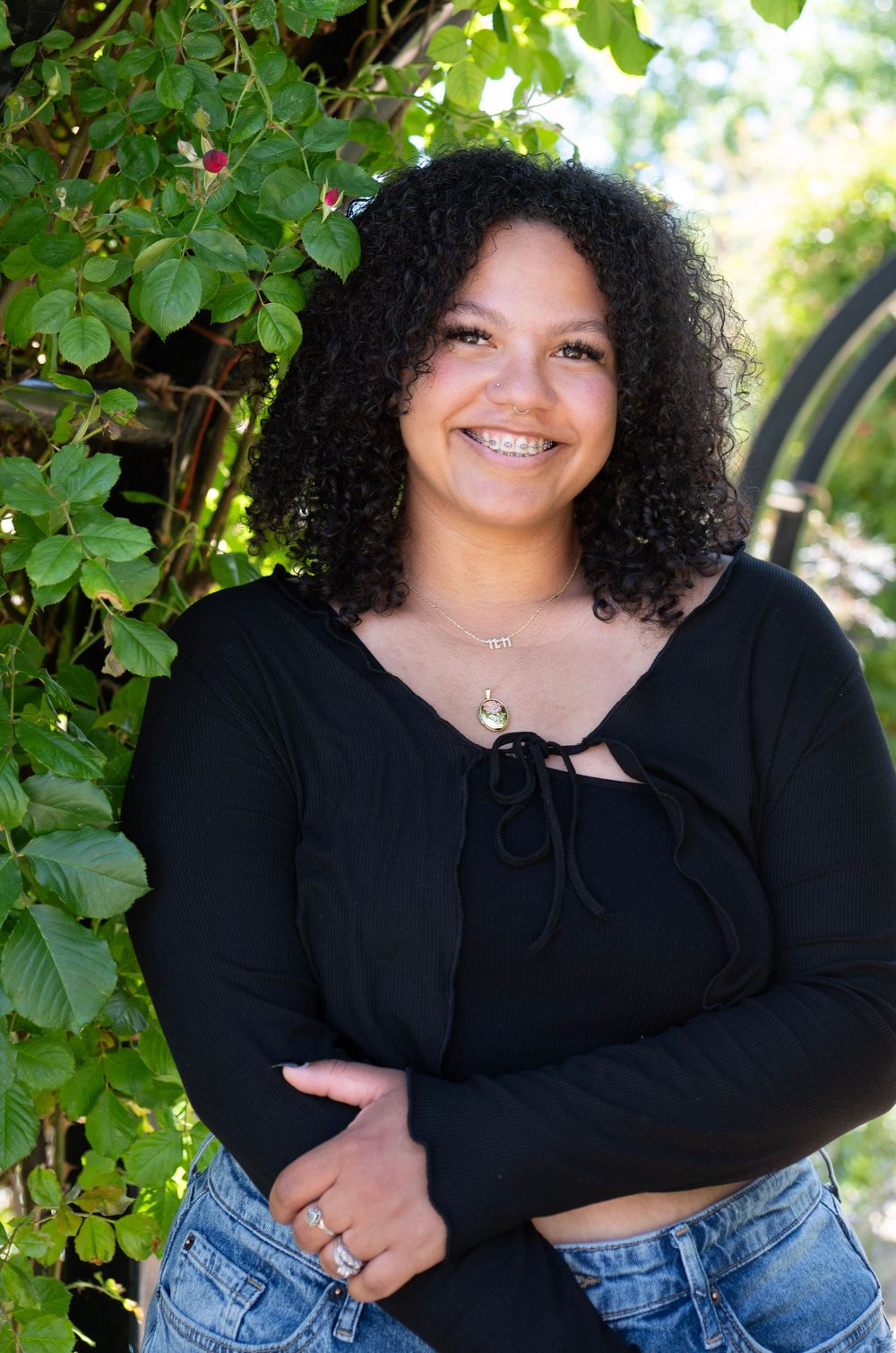 Smiling young woman with curly hair wearing a black top and jeans, posing outdoors near green foliage.