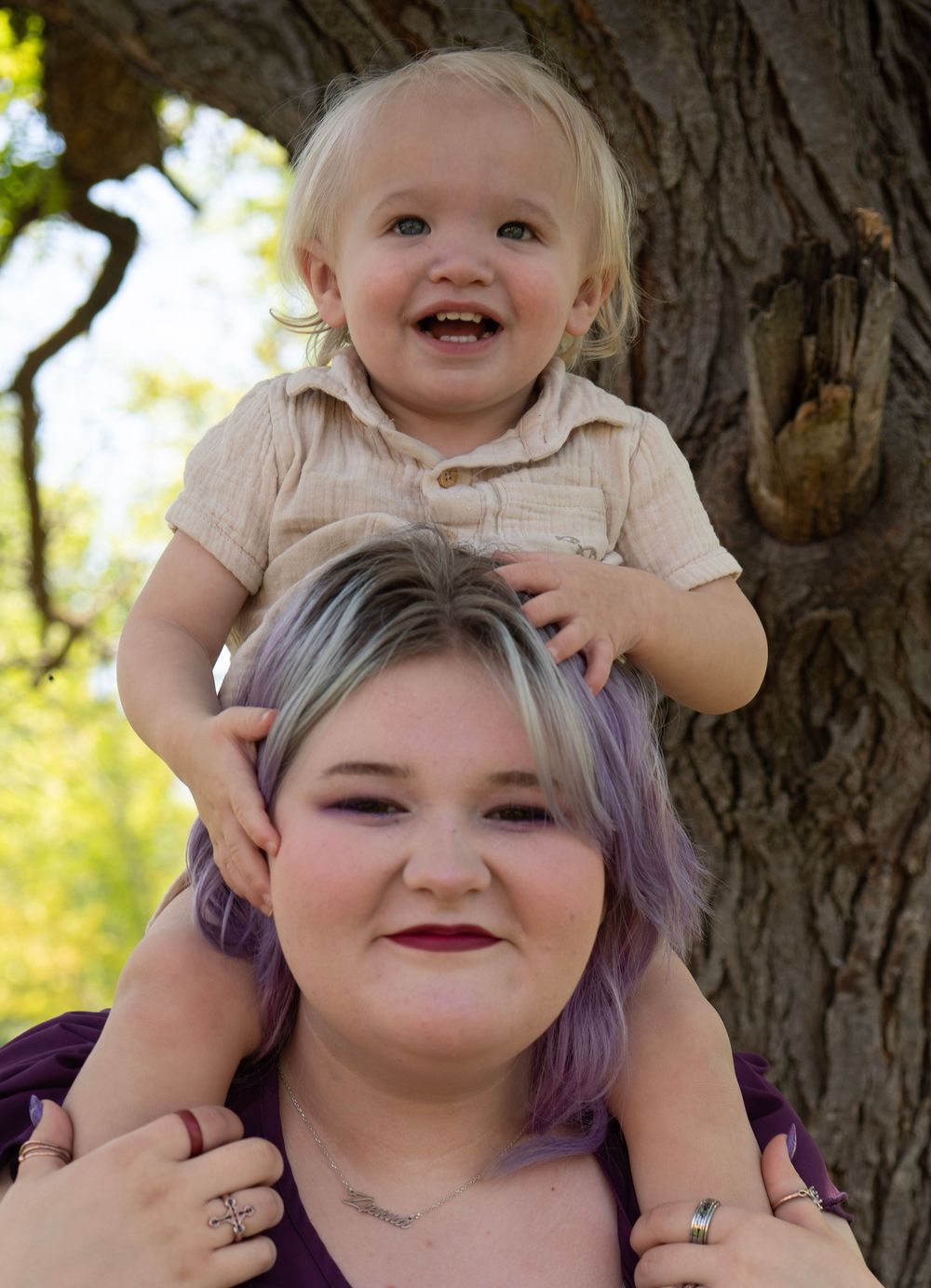 Woman with purple-highlighted hair smiling with a laughing toddler sitting on her shoulders outdoors.