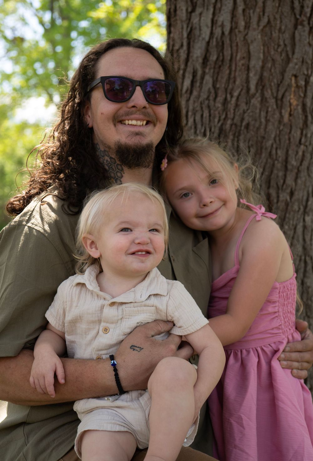 A smiling man with long hair and sunglasses poses with two young children near a tree outdoors.