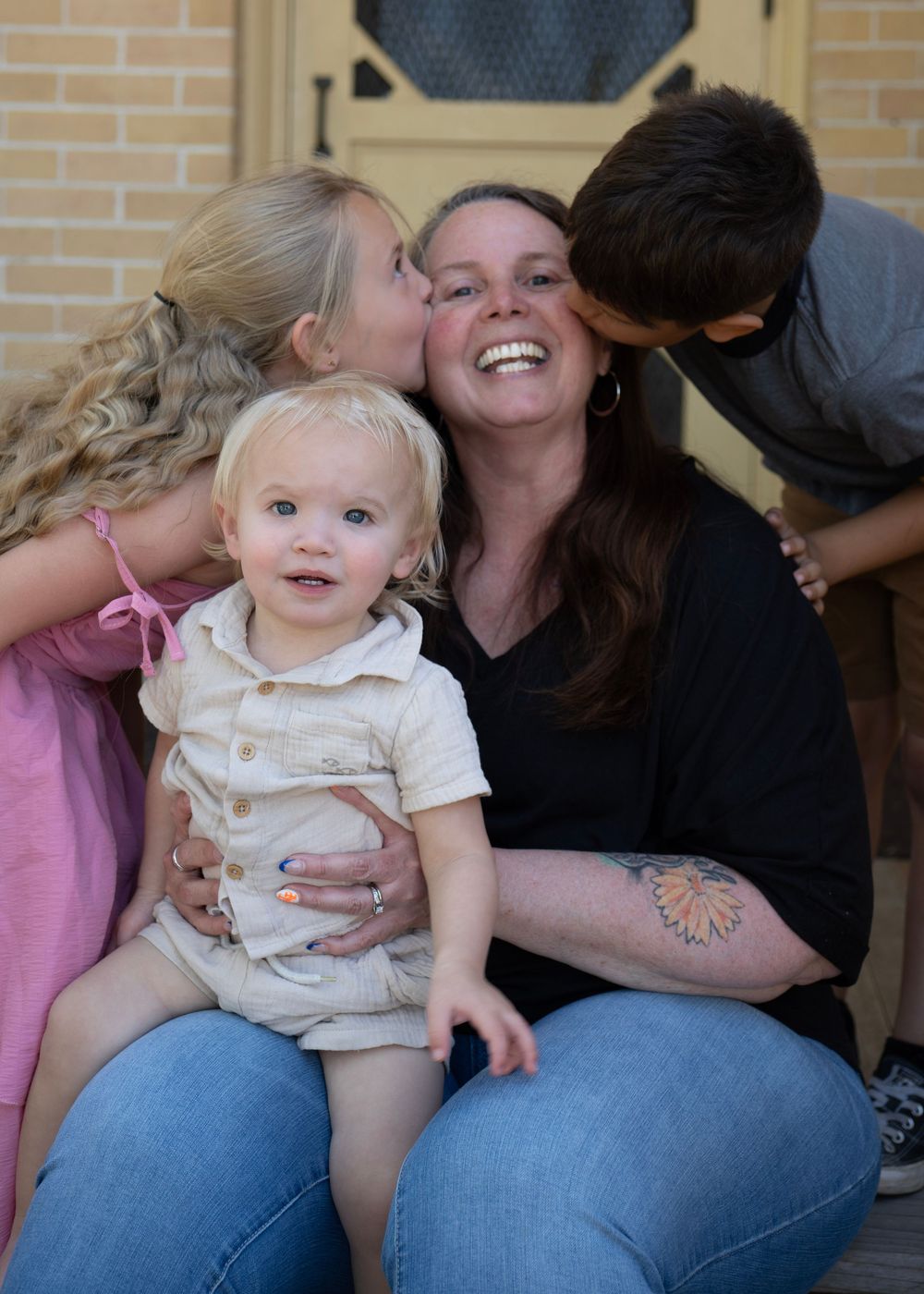 A smiling woman holds a baby while two children kiss her cheeks in a heartwarming family portrait outdoors.