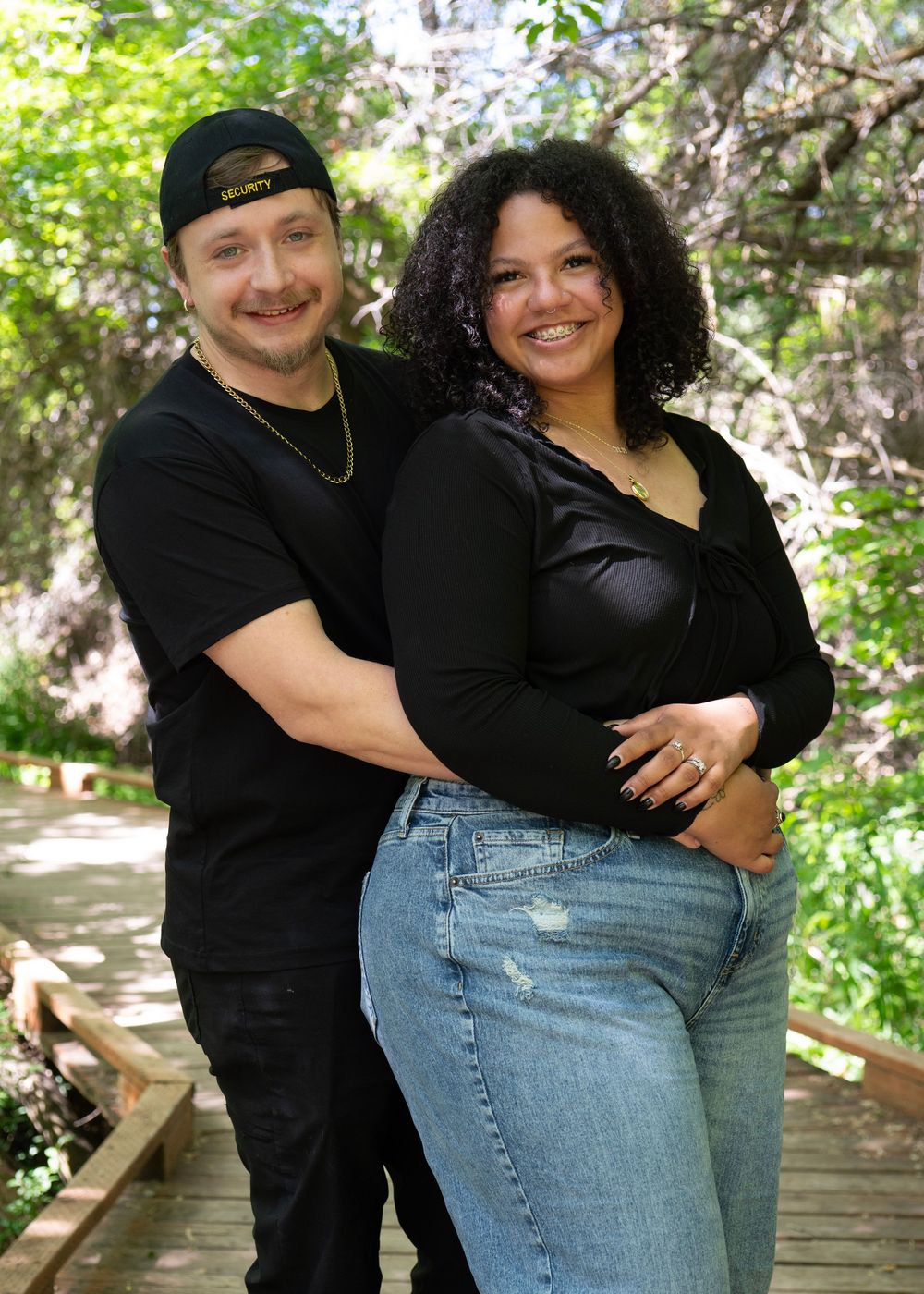 A smiling couple in black outfits poses on a wooden bridge surrounded by lush green trees in an outdoor setting.