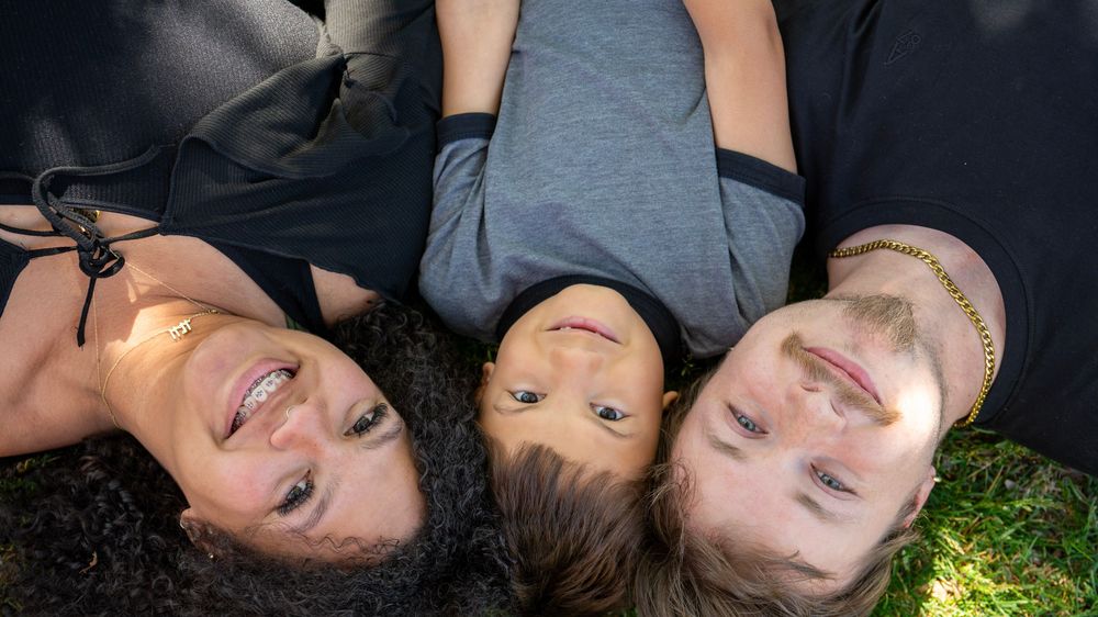 Three friends lying on grass looking up smiling, captured from above in a cheerful outdoor portrait.