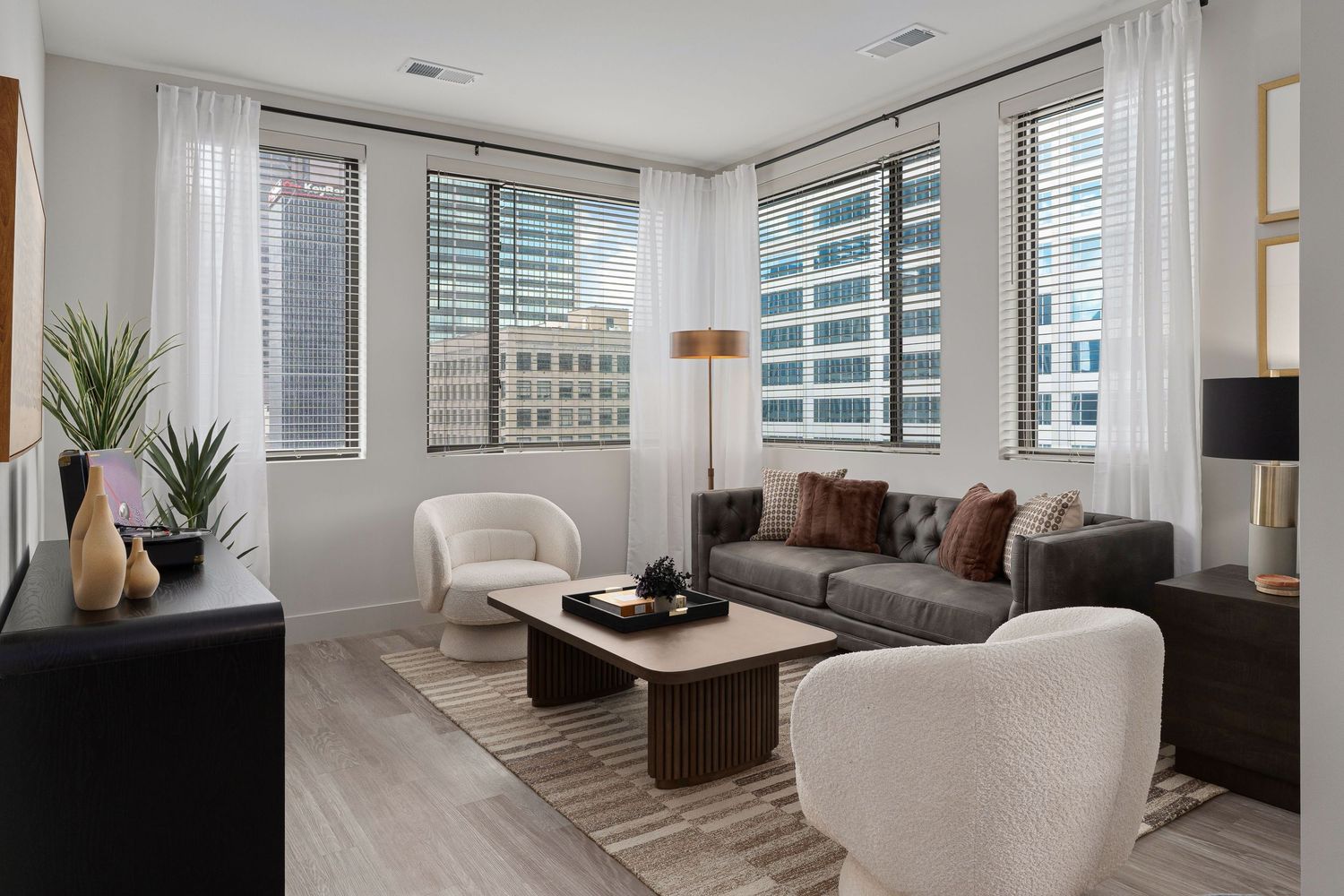 Model apartment with modern living room with grey sofa, white accent chairs, hardwood floors and large windows overlooking urban skyline.
