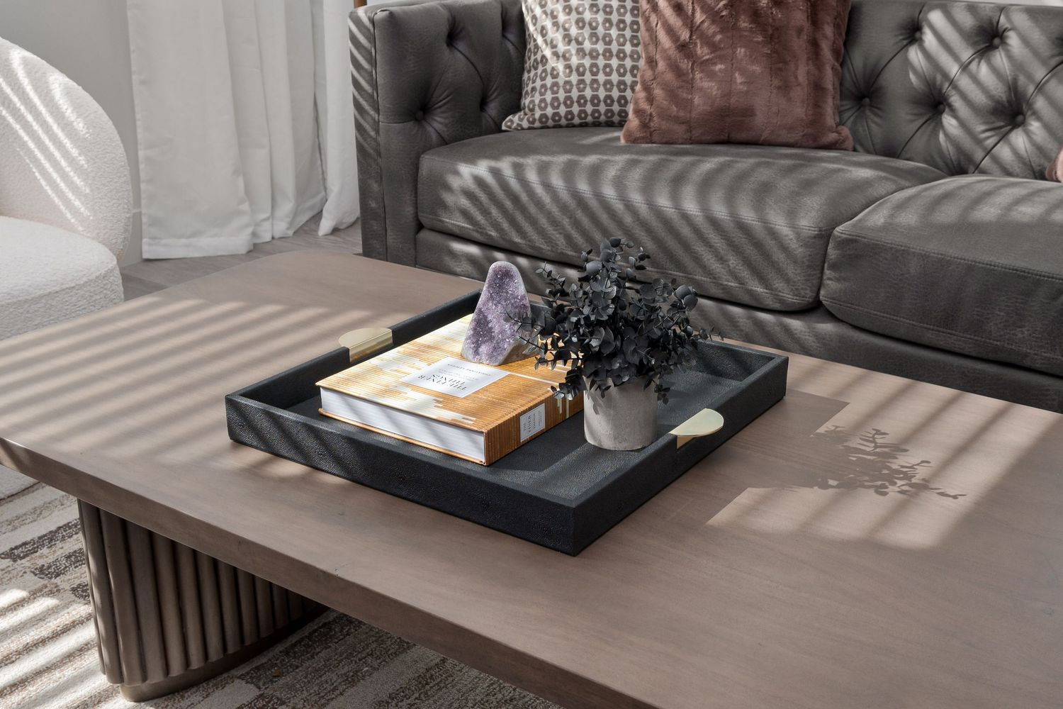 Detail photo of a modern coffee table with decorative tray displaying books and purple flowers, casting striped shadows in sunlight.