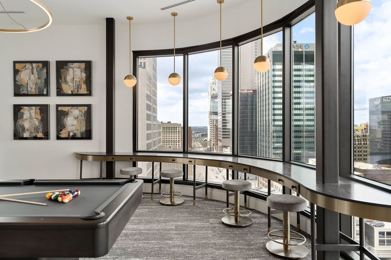Modern game room with pool table, curved windows, city views, bar seating, and pendant lighting in an upscale high-rise apartment.