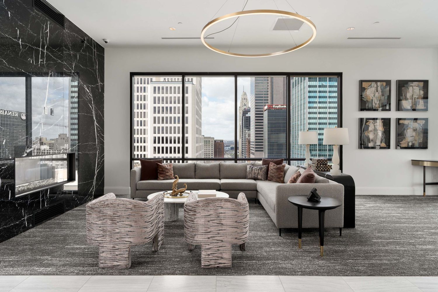 A modern luxury apartment club room with grey sectional sofa, marble coffee tables and floor-to-ceiling windows overlooking a city skyline.