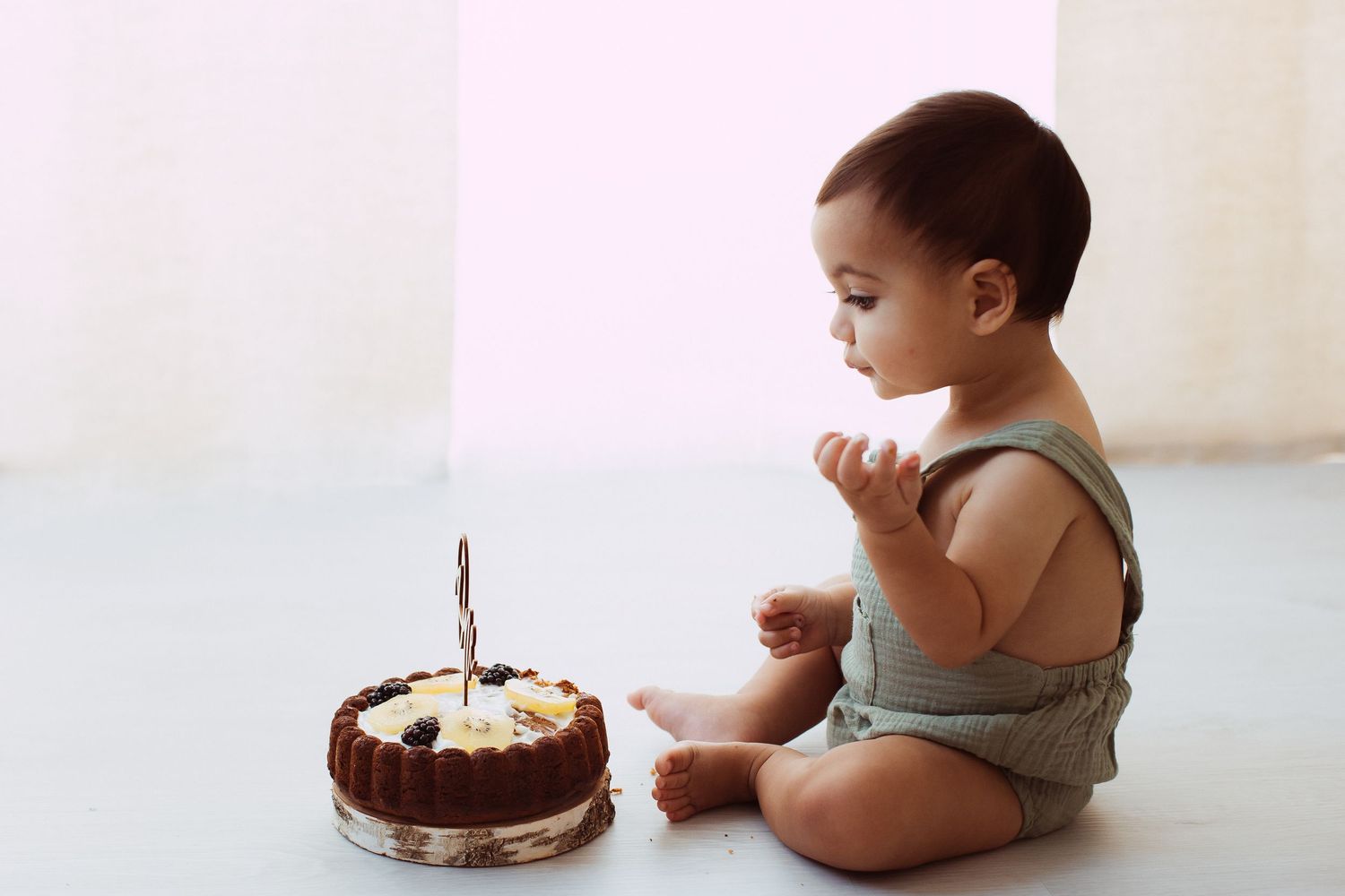 Baby in grey onesie sitting beside chocolate birthday cake on white backdrop.