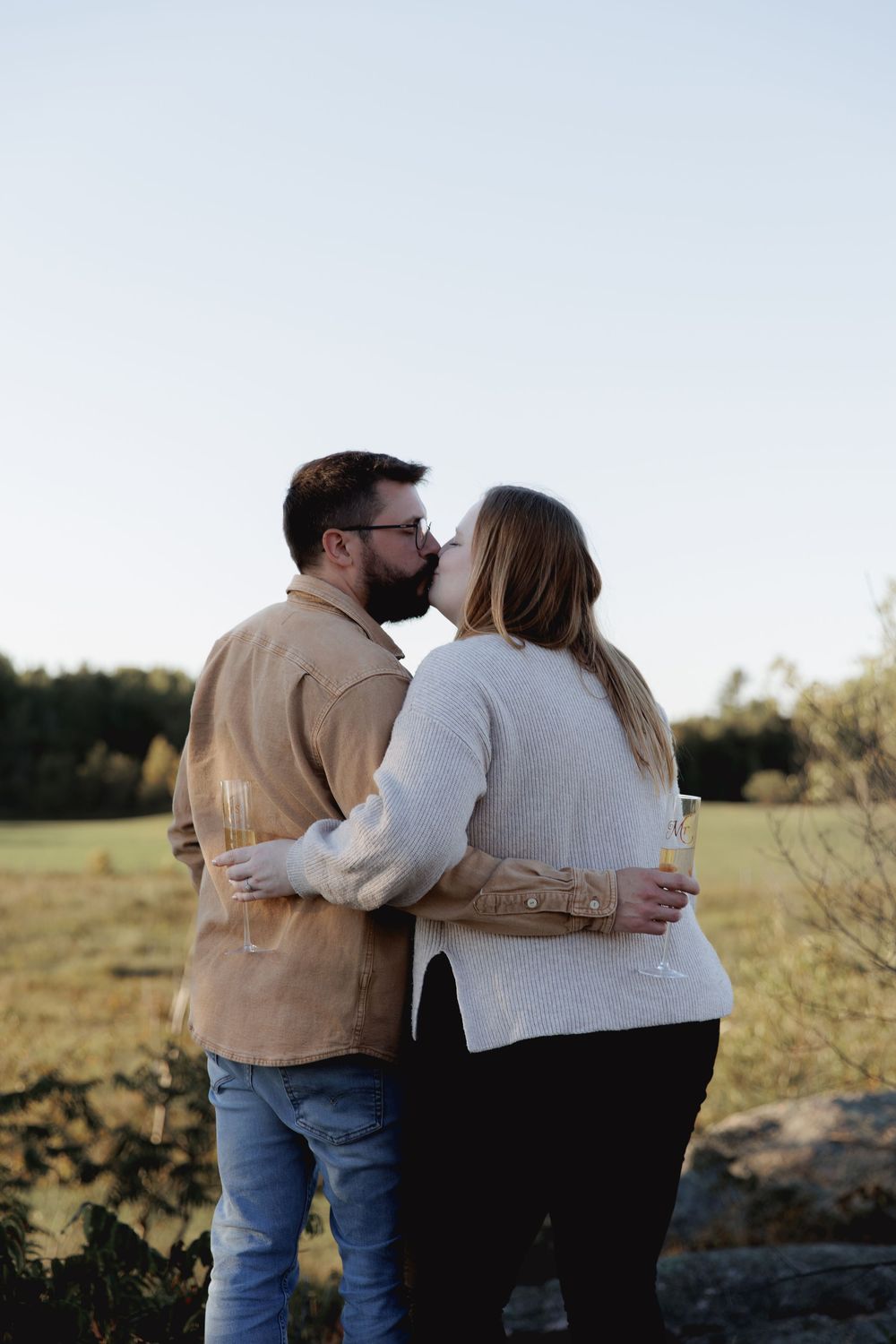 A couple in casual attire embraces while sitting on rocks in a scenic field during golden hour.