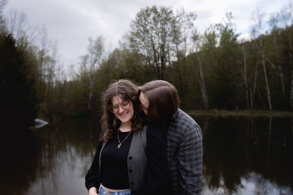 A romantic moment by a serene lake surrounded by trees on an overcast day.