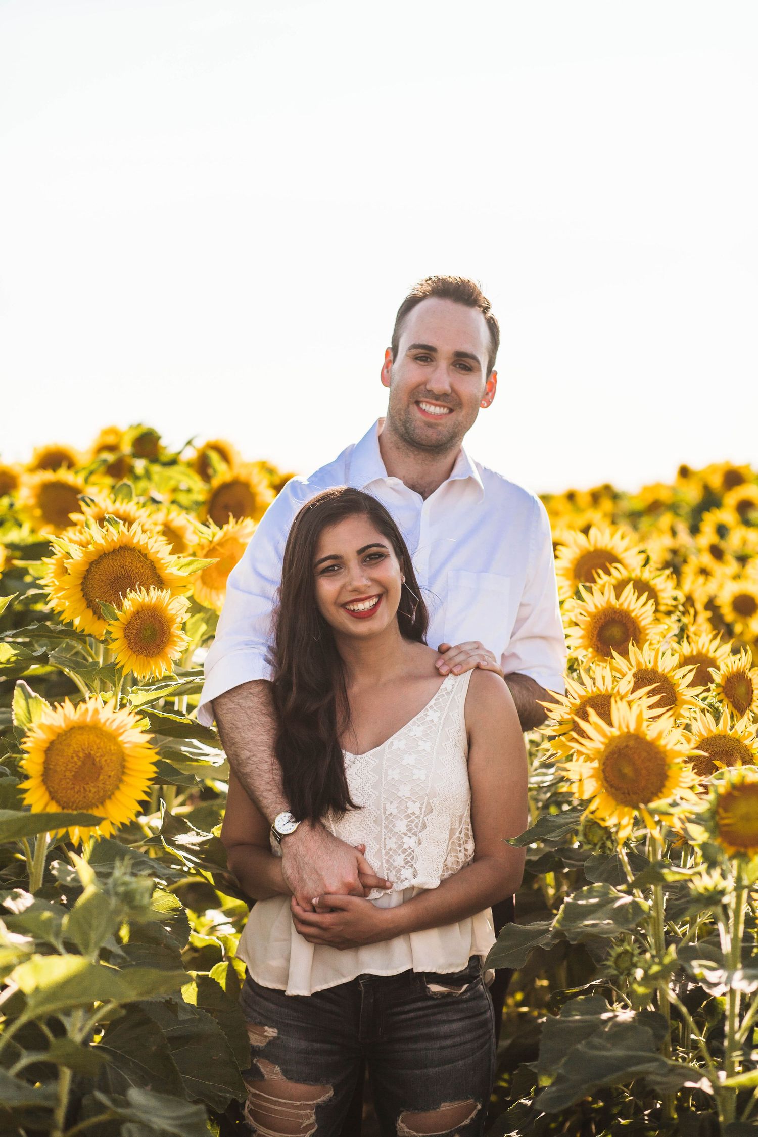 A man and woman wearing white smiling surrounded by a field of sunflowers.