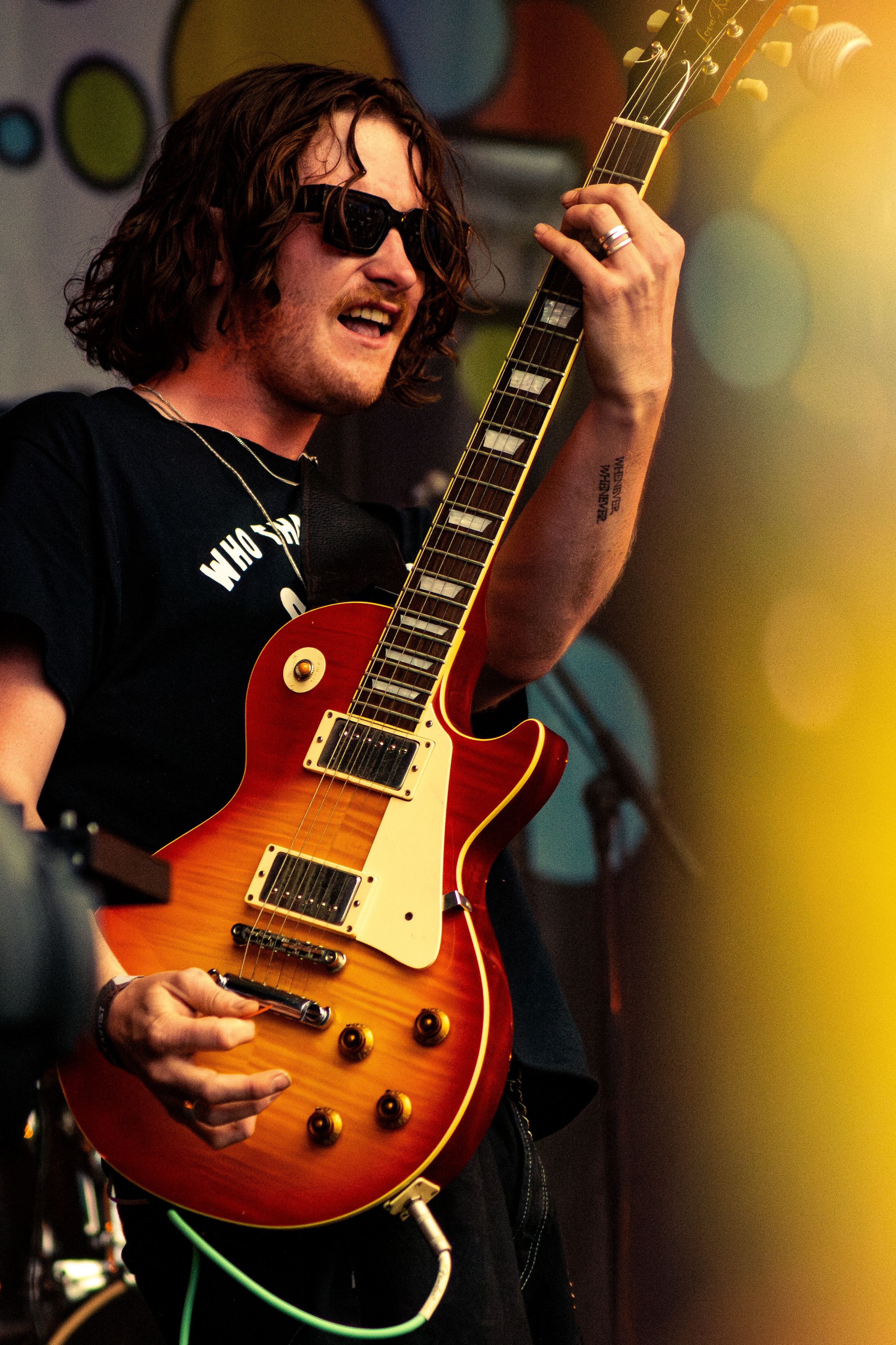 Guitarist plays sunburst Les Paul guitar on colorful stage wearing sunglasses and dark shirt.