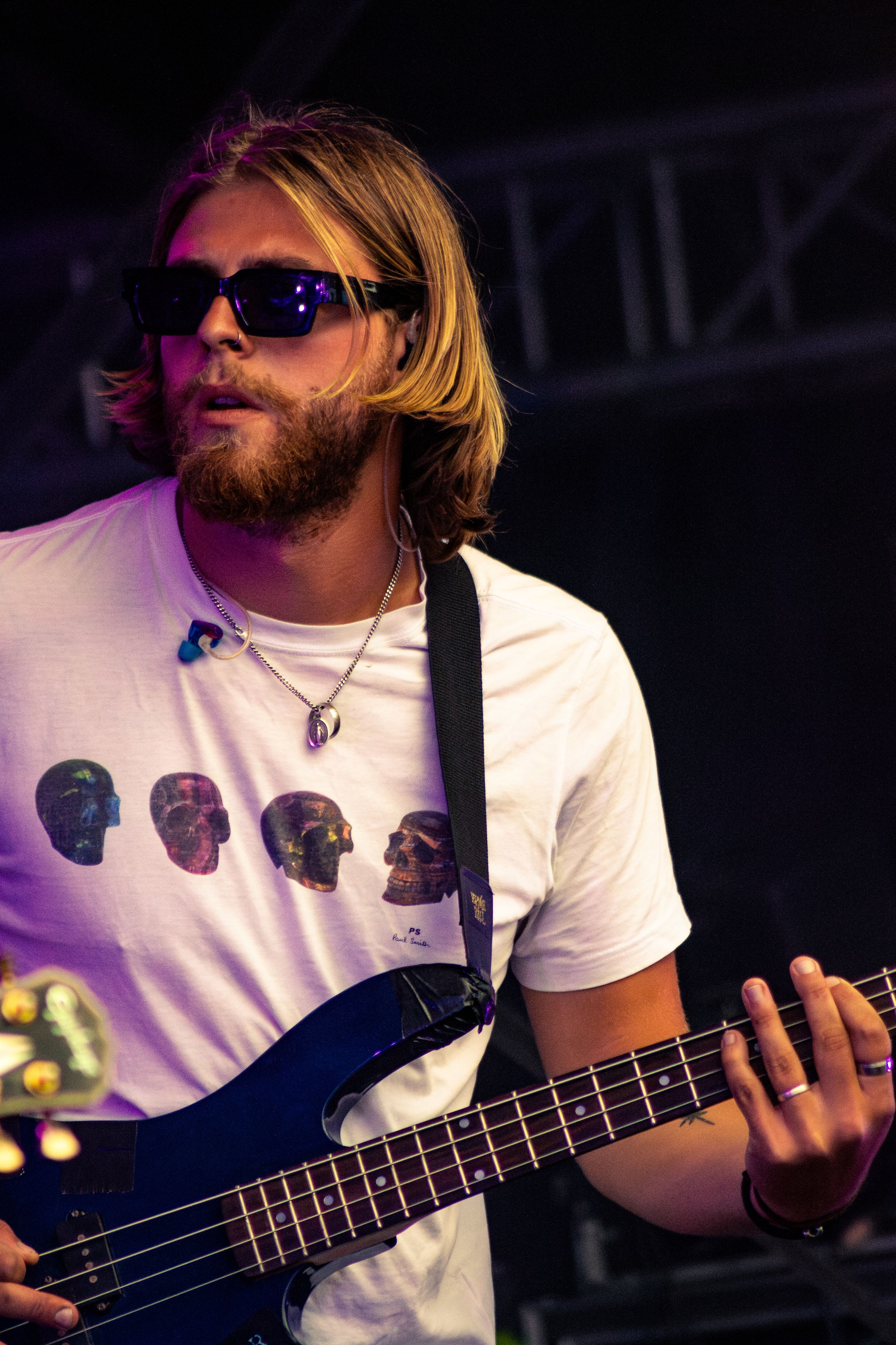 Bass guitarist performing on stage wearing sunglasses and a white t-shirt with skull designs under purple lighting.