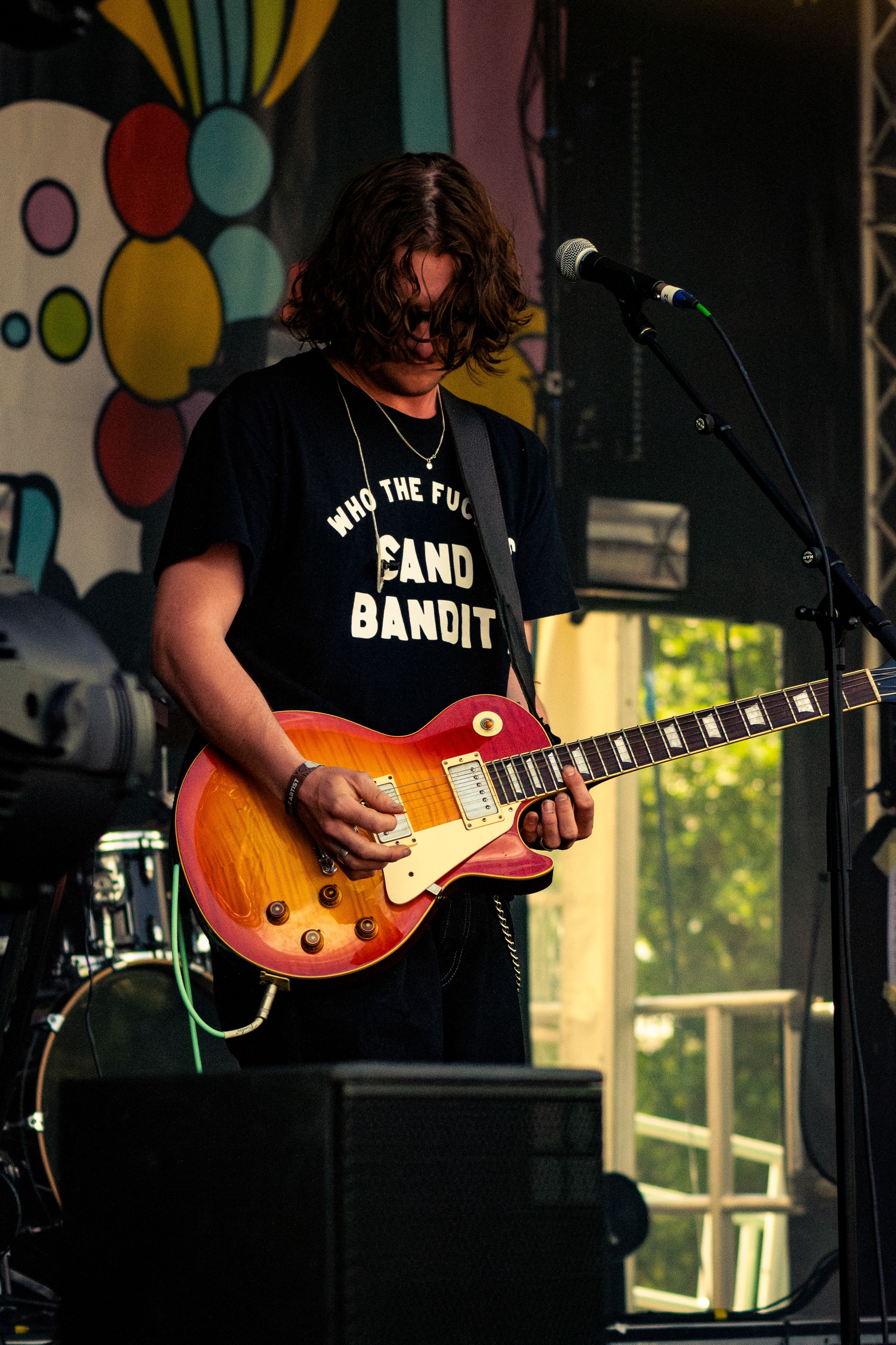 Guitarist in a Sand Bandit t-shirt playing a sunburst Les Paul guitar against a colorful backdrop.