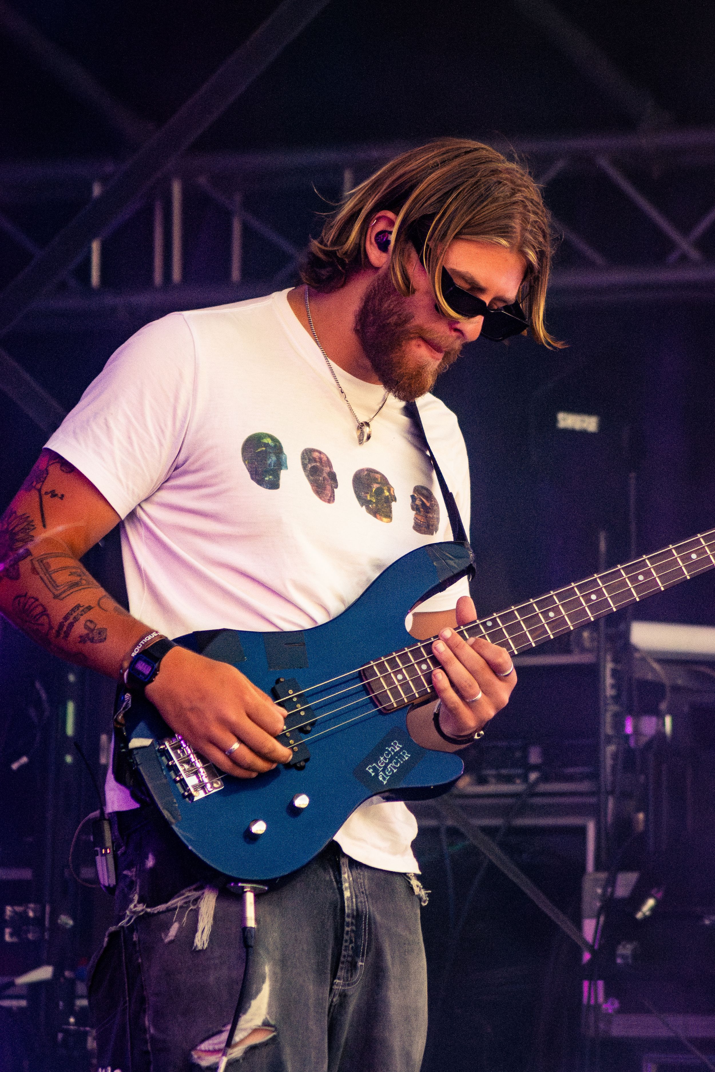 Musician playing blue bass guitar wearing skull t-shirt and sunglasses on outdoor festival stage.