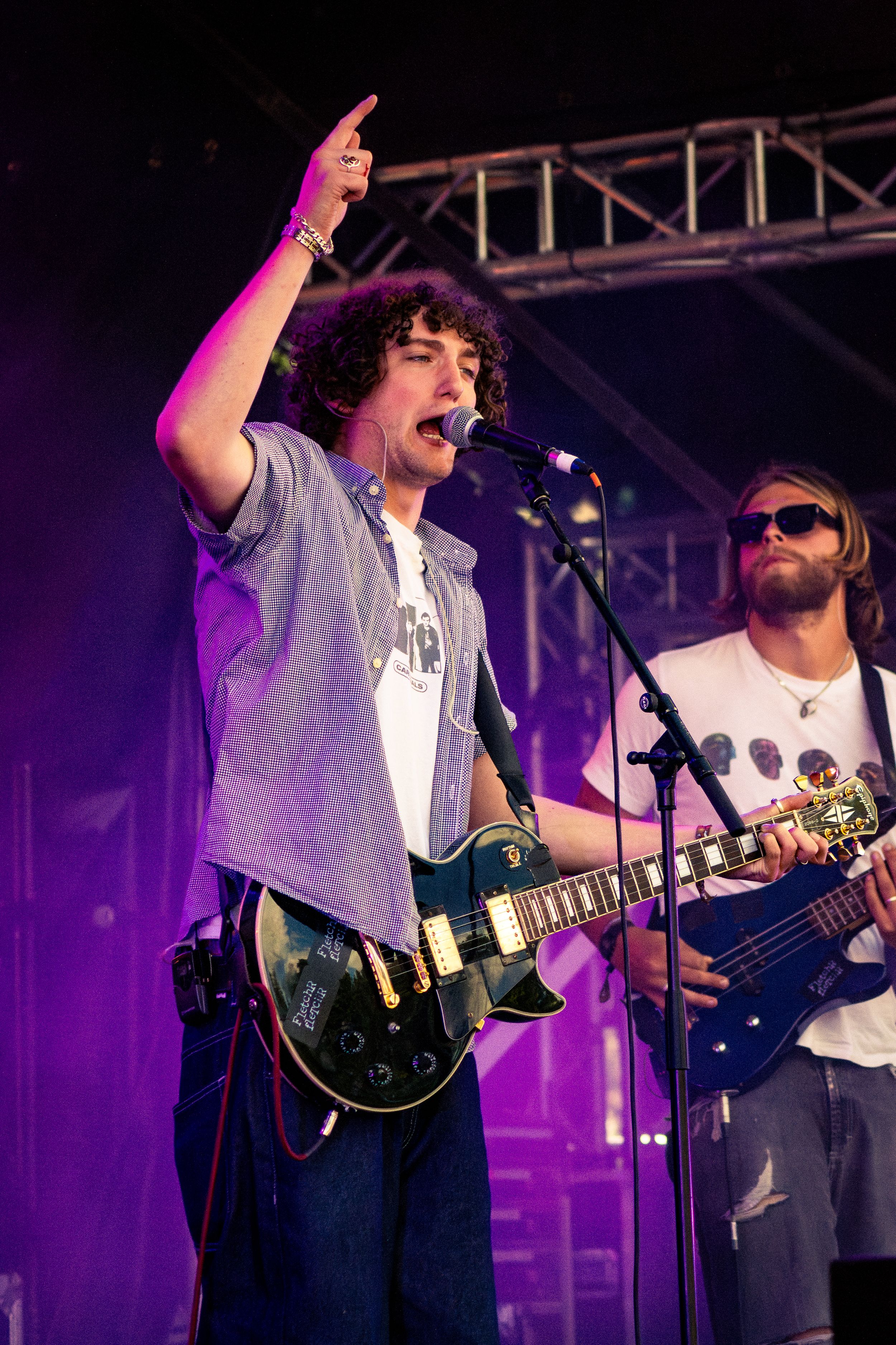 Rock band members performing on stage with guitars under purple lighting and metal truss structures.