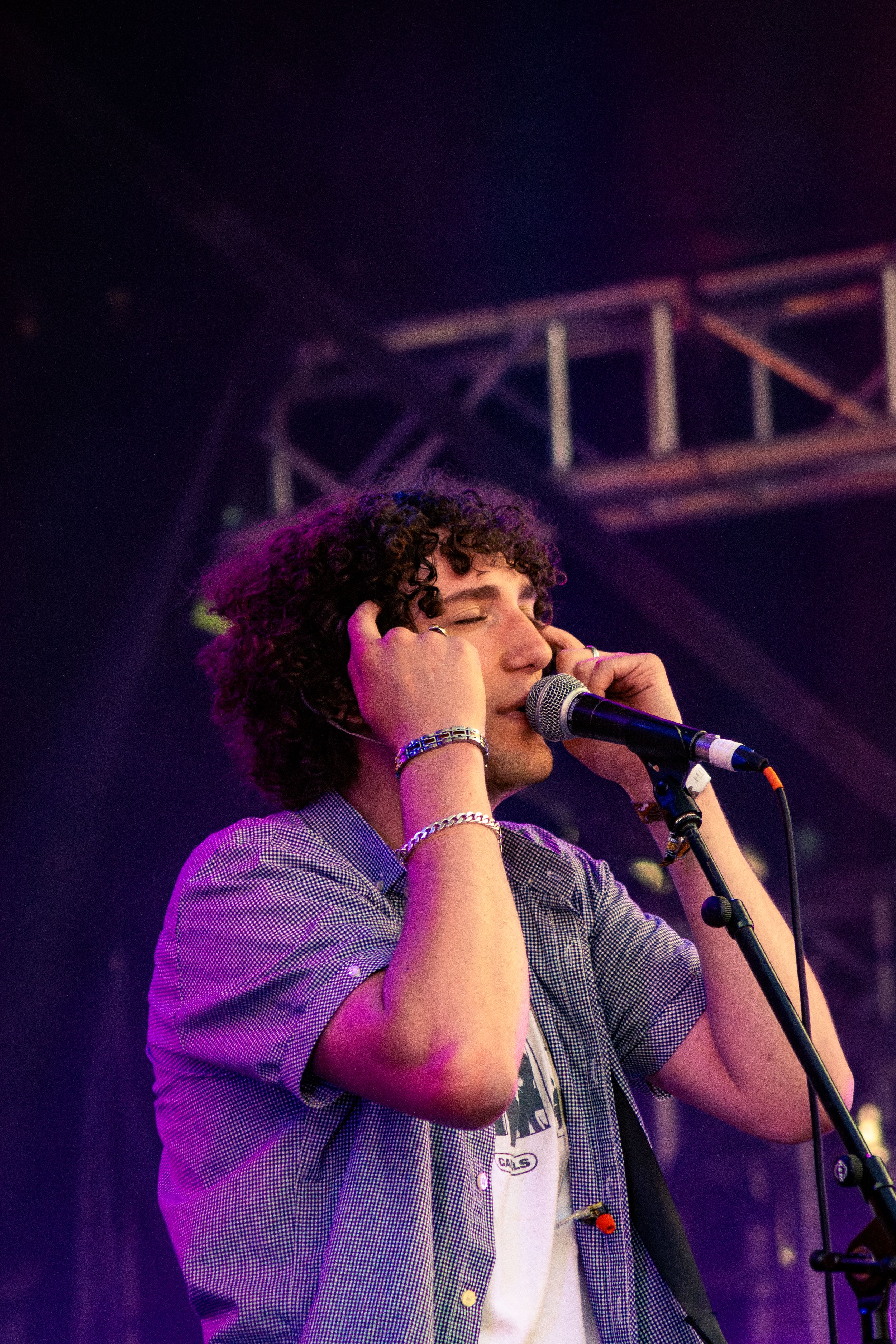 Vocalist singing into microphone wearing checkered shirt with bracelets under stage lights.