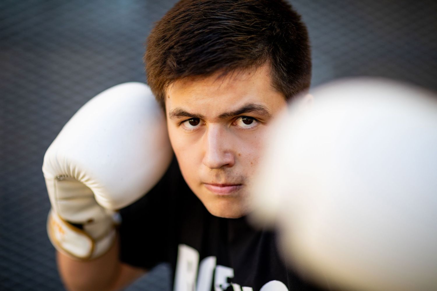 A high school senior in a black t-shirt holds up defensive gloves against a dark background with focus on white boxing gloves.