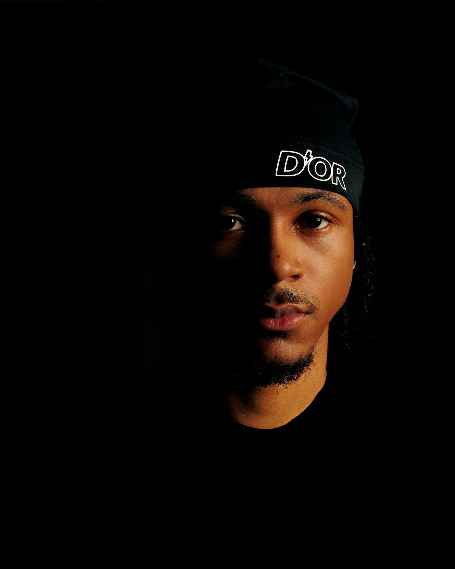 A dramatic portrait in dark lighting shows someone wearing a black Dior headband against a black background.