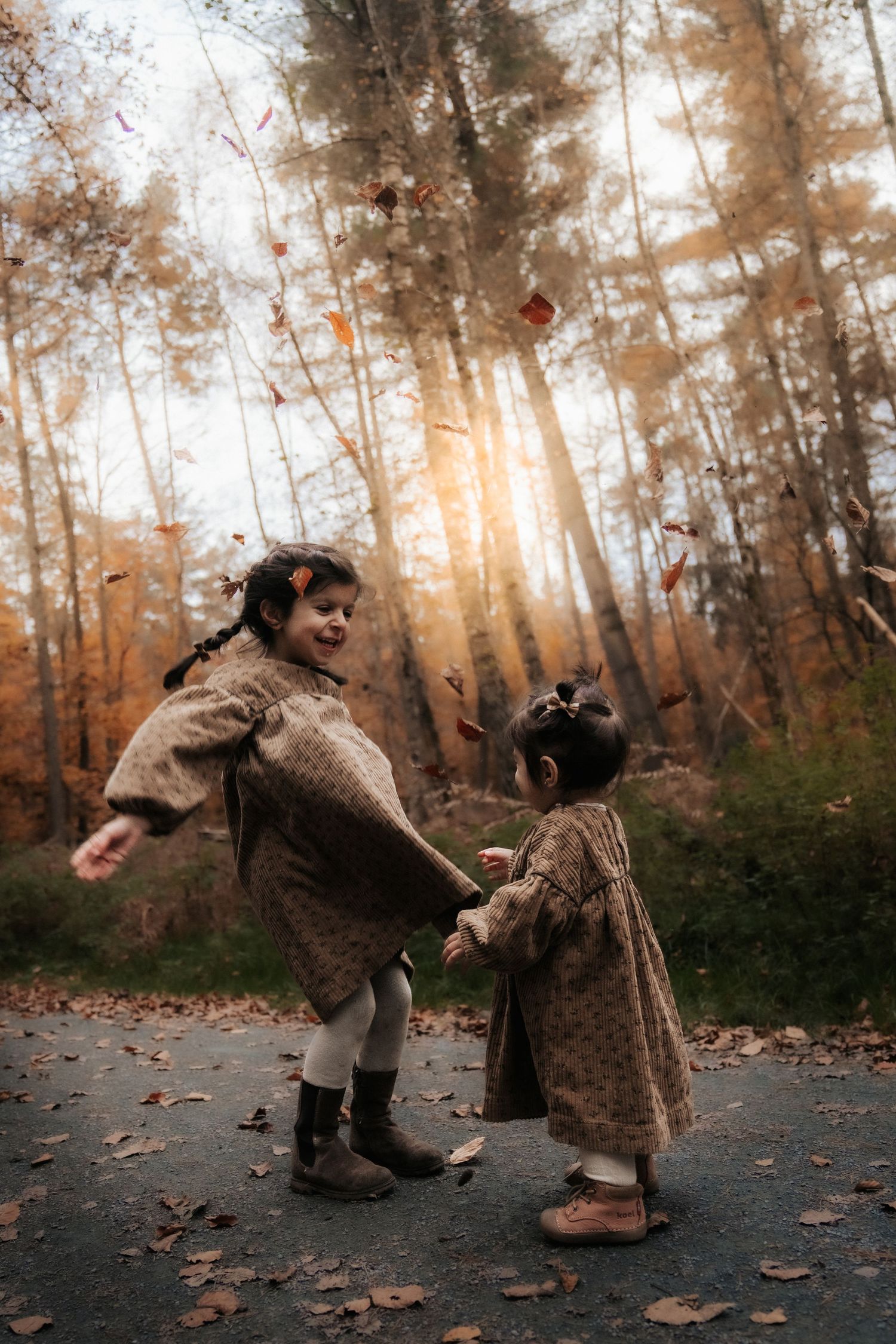 Children in vintage-style clothing dance together on a forest path during autumn as sunlight filters through the trees.