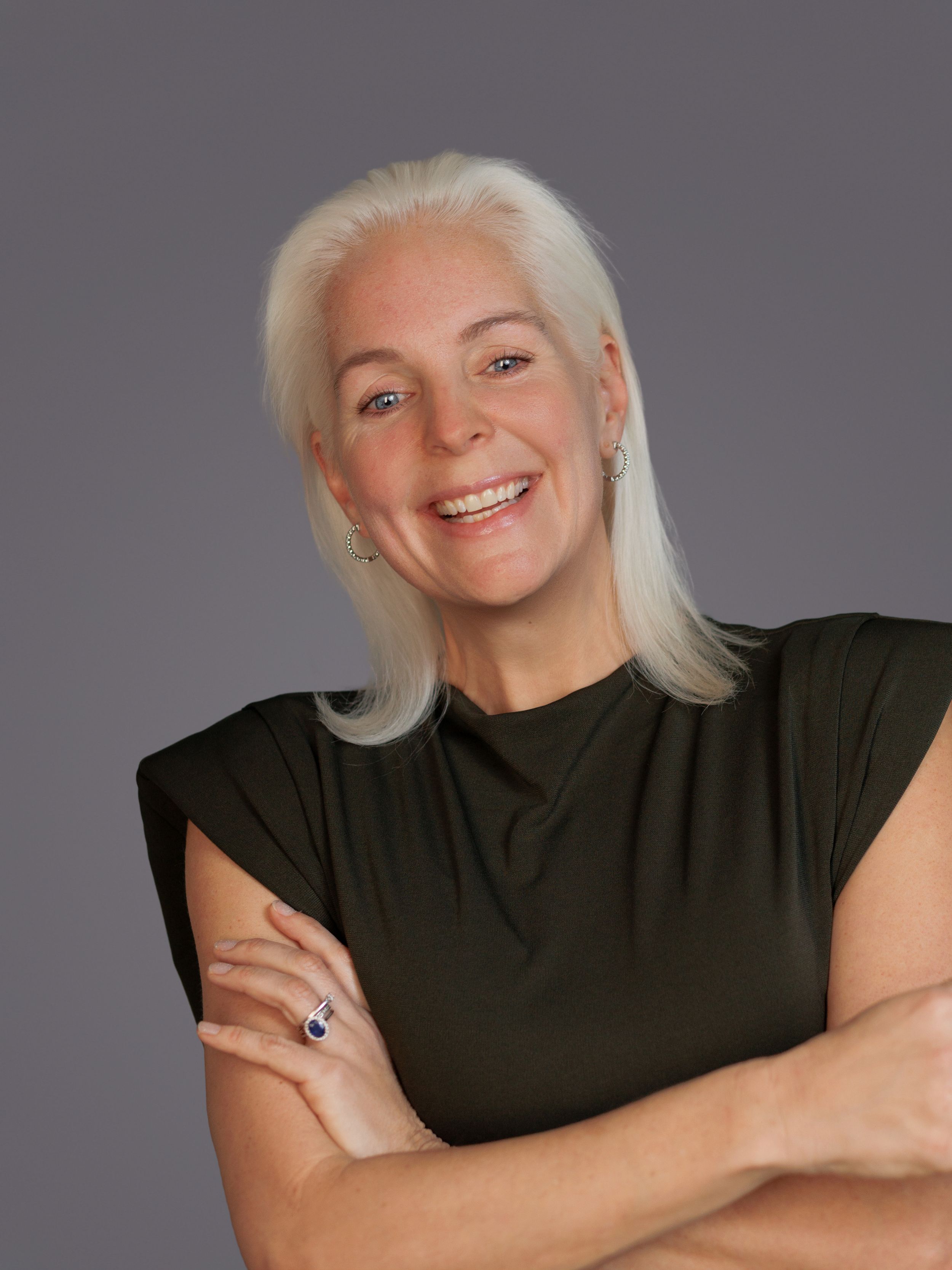 Smiling woman with white hair wearing a dark green sleeveless top, arms crossed, posing against a gray background.
