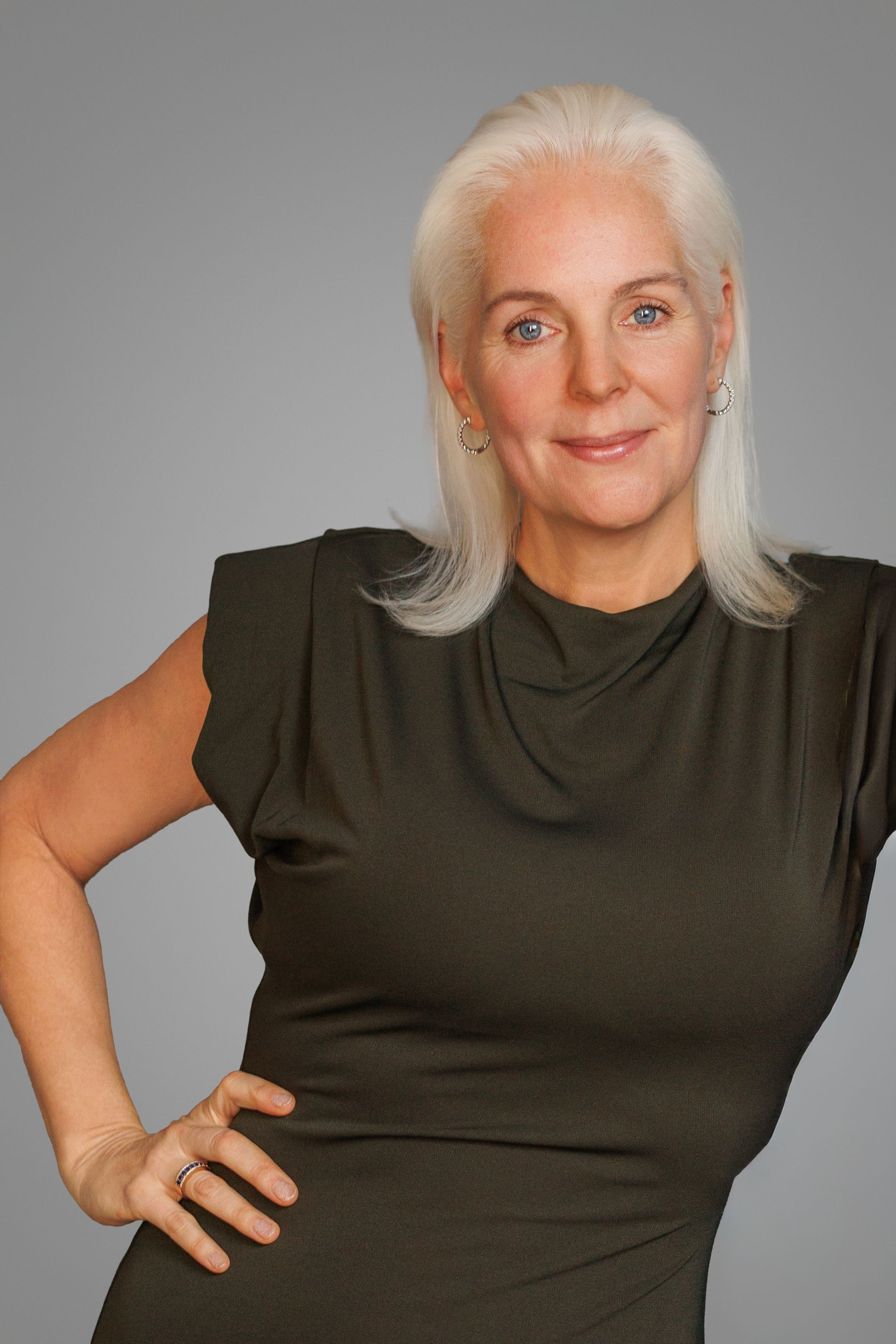 Confident mature woman with silver hair in dark olive sleeveless dress posing with hand on hip against gray background.