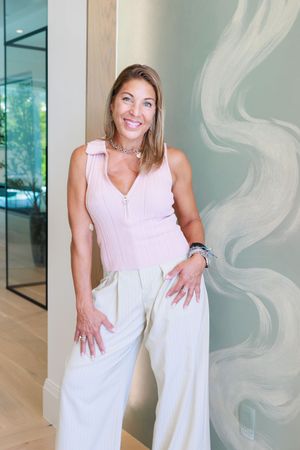 Smiling brunette woman in pink top and white wide-leg pants posing against decorative wall.