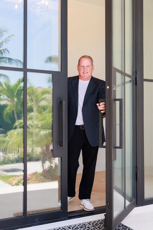 Smiling man in dark blazer and white shirt opening glass door with palm trees visible outside.