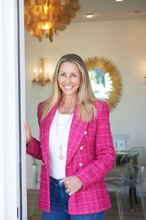 Blonde woman in pink tweed blazer smiles while leaning against doorframe in elegant interior space.
