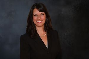 Professional woman in black blazer and pearl necklace smiling against dark studio background.