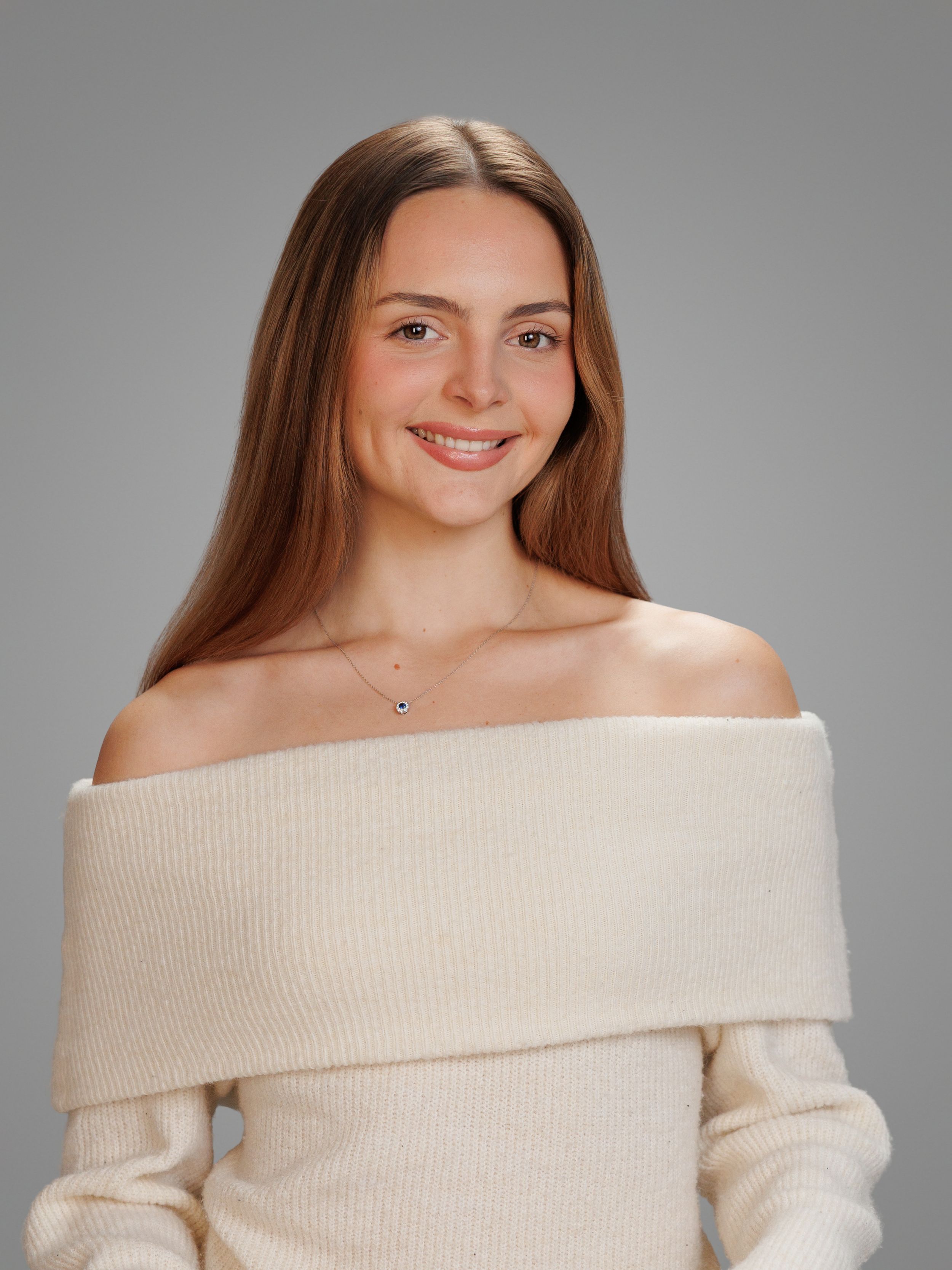 Smiling young woman with long brown hair wearing an off-shoulder cream knit sweater against a gray background.