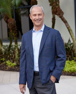 Professional man in navy blazer smiling outdoors near tropical palm trees in a business portrait photo.