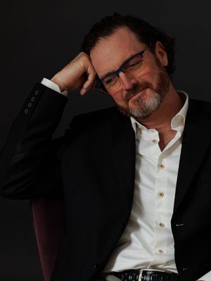 Middle-aged man with glasses and beard in black blazer, eyes closed, resting head on hand against dark background.