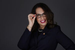 Professional woman in navy blazer smiling, adjusting glasses, wearing pearl necklace, dark studio background.