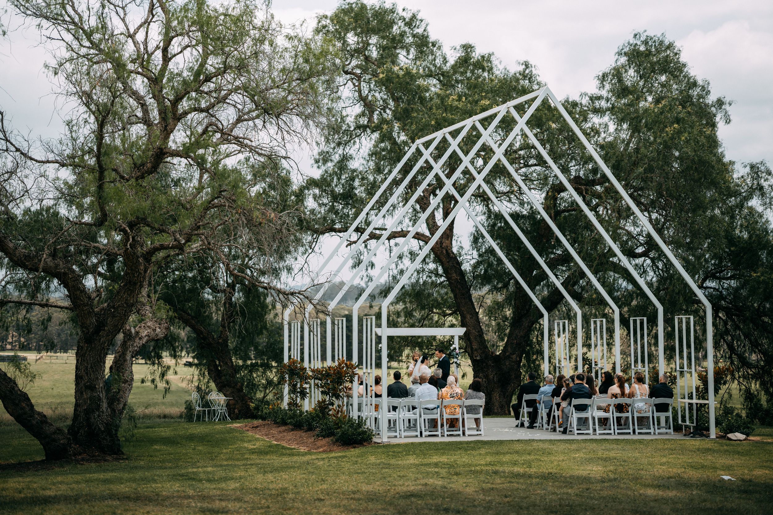 Modern glass greenhouse wedding venue with white frame structure surrounded by trees.