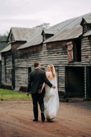 Couple walks hand in hand past rustic wooden barns on a dirt path.