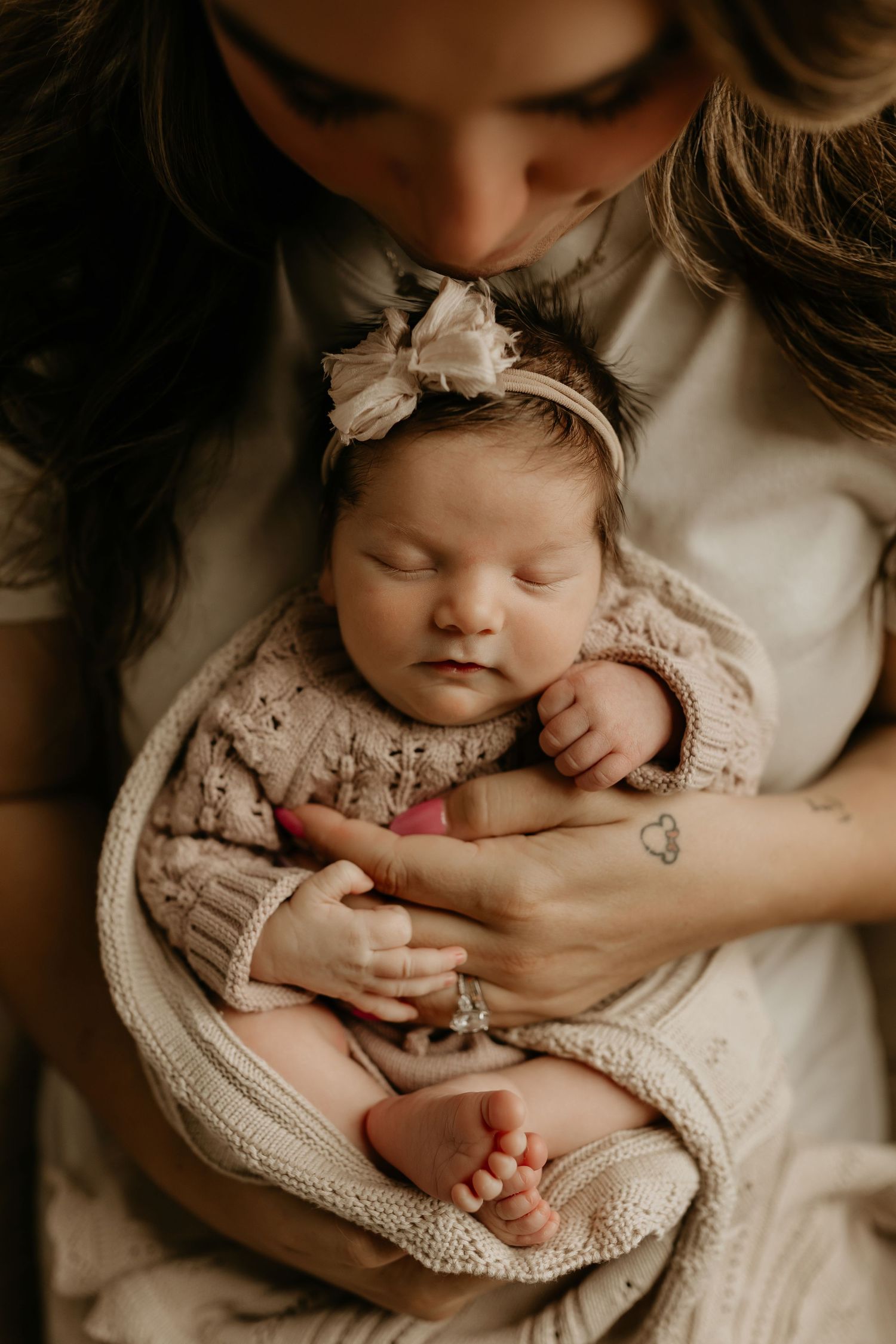 A sleeping newborn baby wearing a knit romper and bow headband is cradled in loving arms against soft fabric. Tulsa newborn photographer