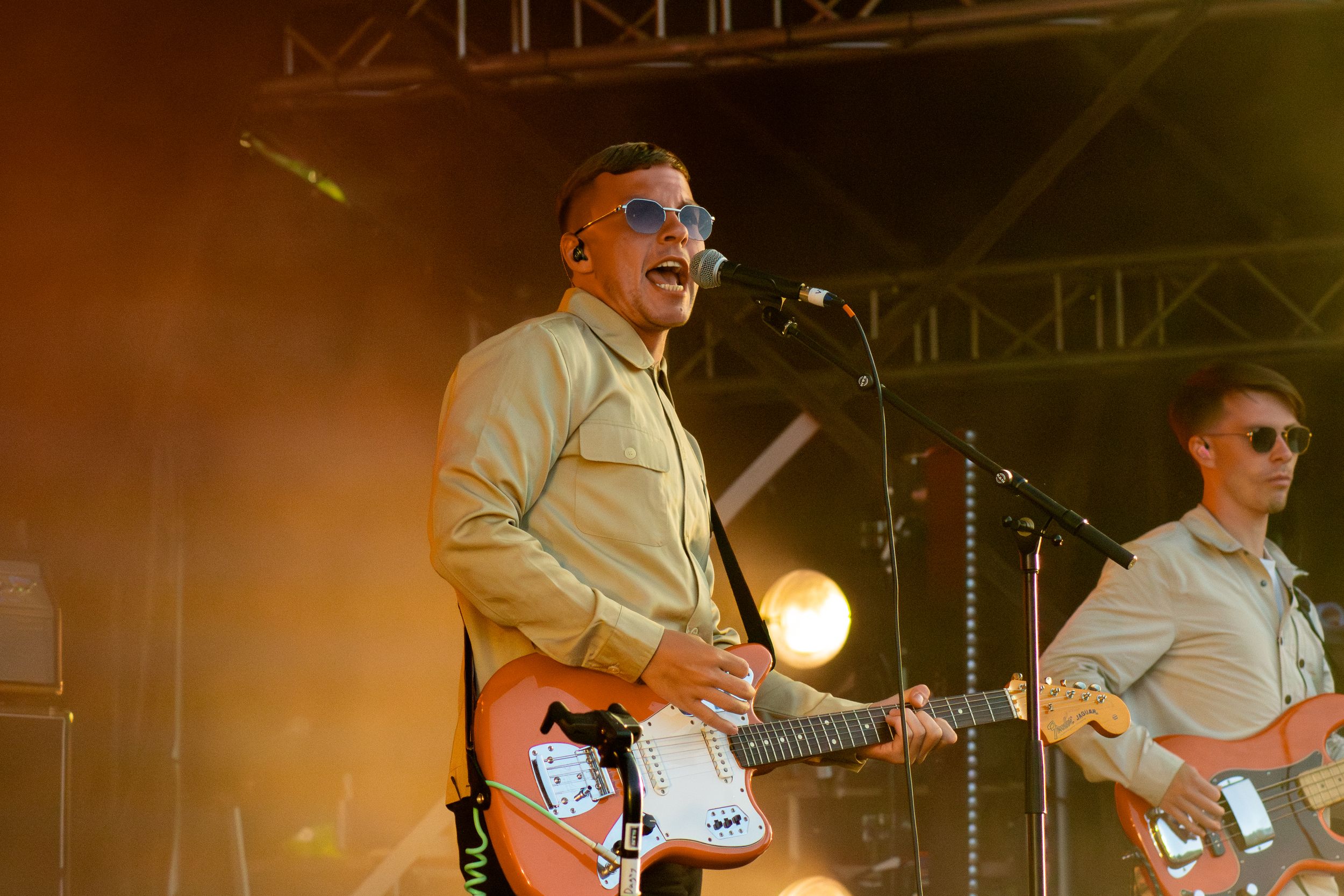 Musicians perform on stage in beige shirts with guitars under dramatic concert lighting.