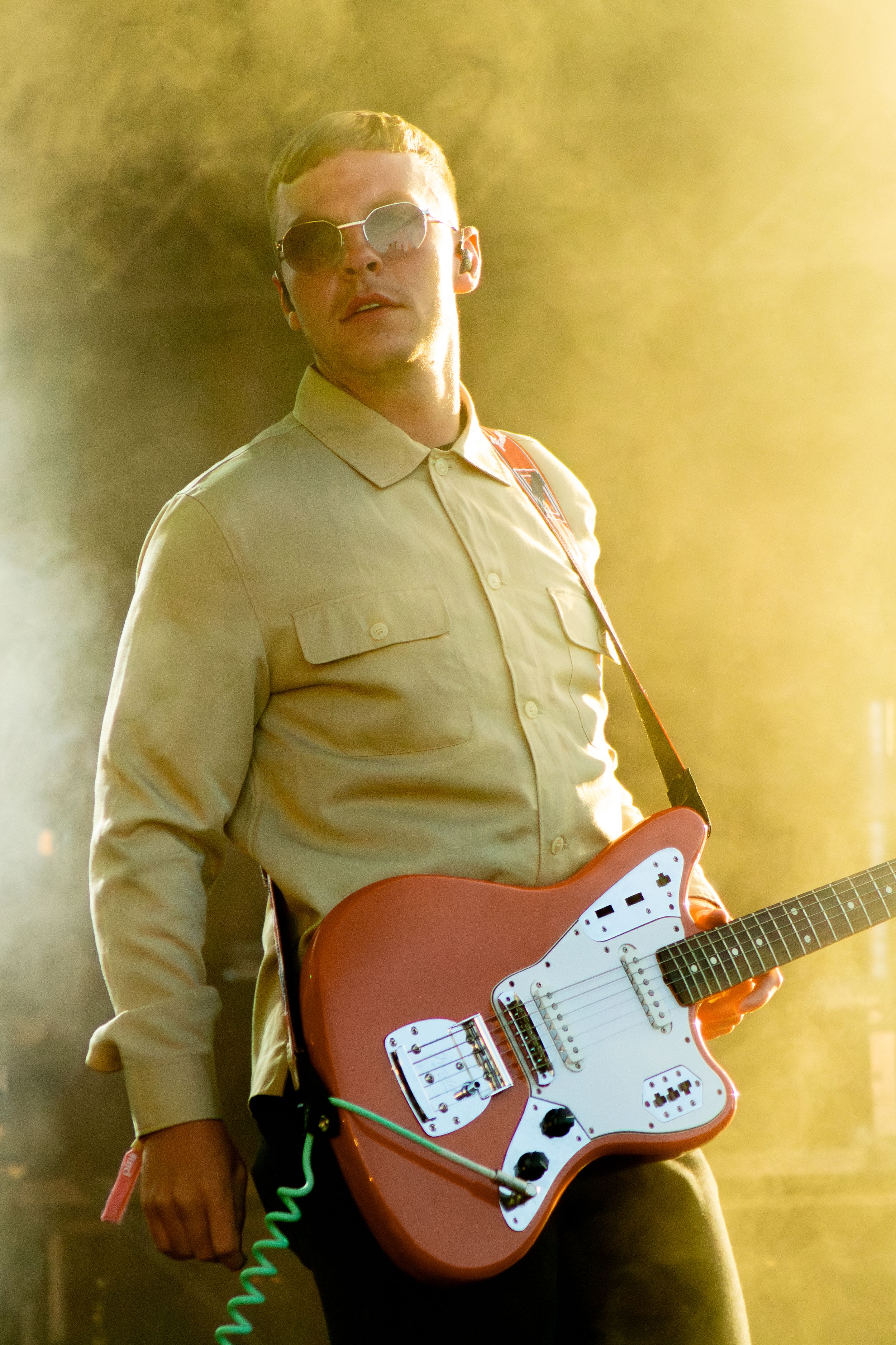 Guitarist in beige button-up shirt performs with orange Jaguar-style electric guitar in golden lighting.
