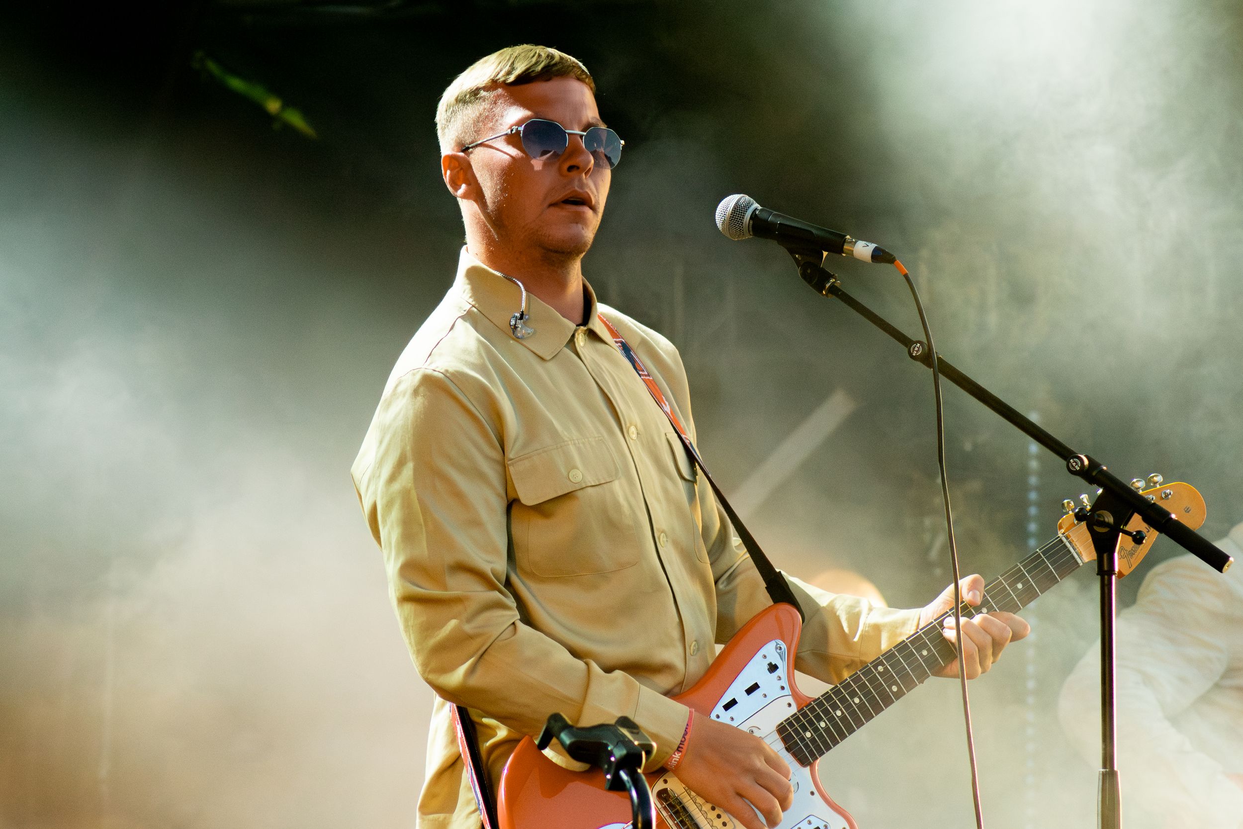 Musician in sunglasses plays guitar while singing into microphone on smoky concert stage.