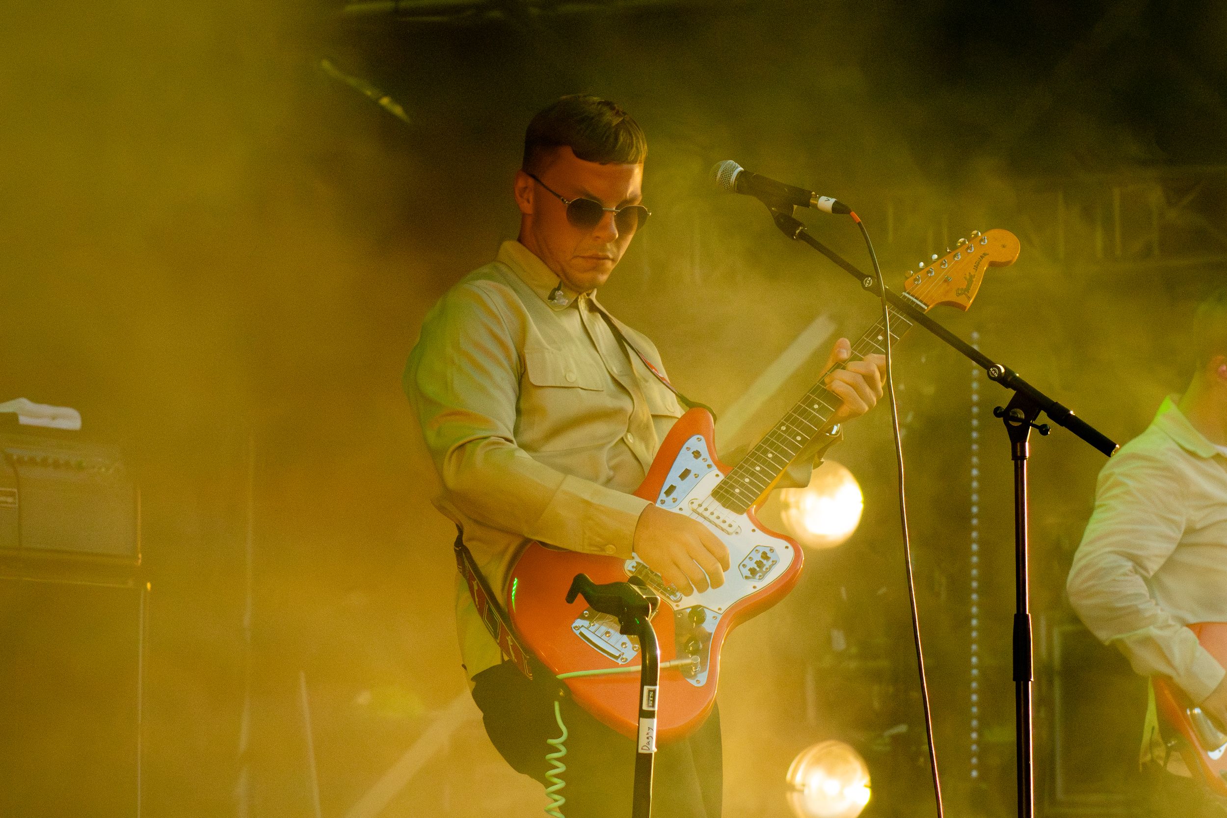 Musician plays orange guitar on stage bathed in warm yellow concert lighting.