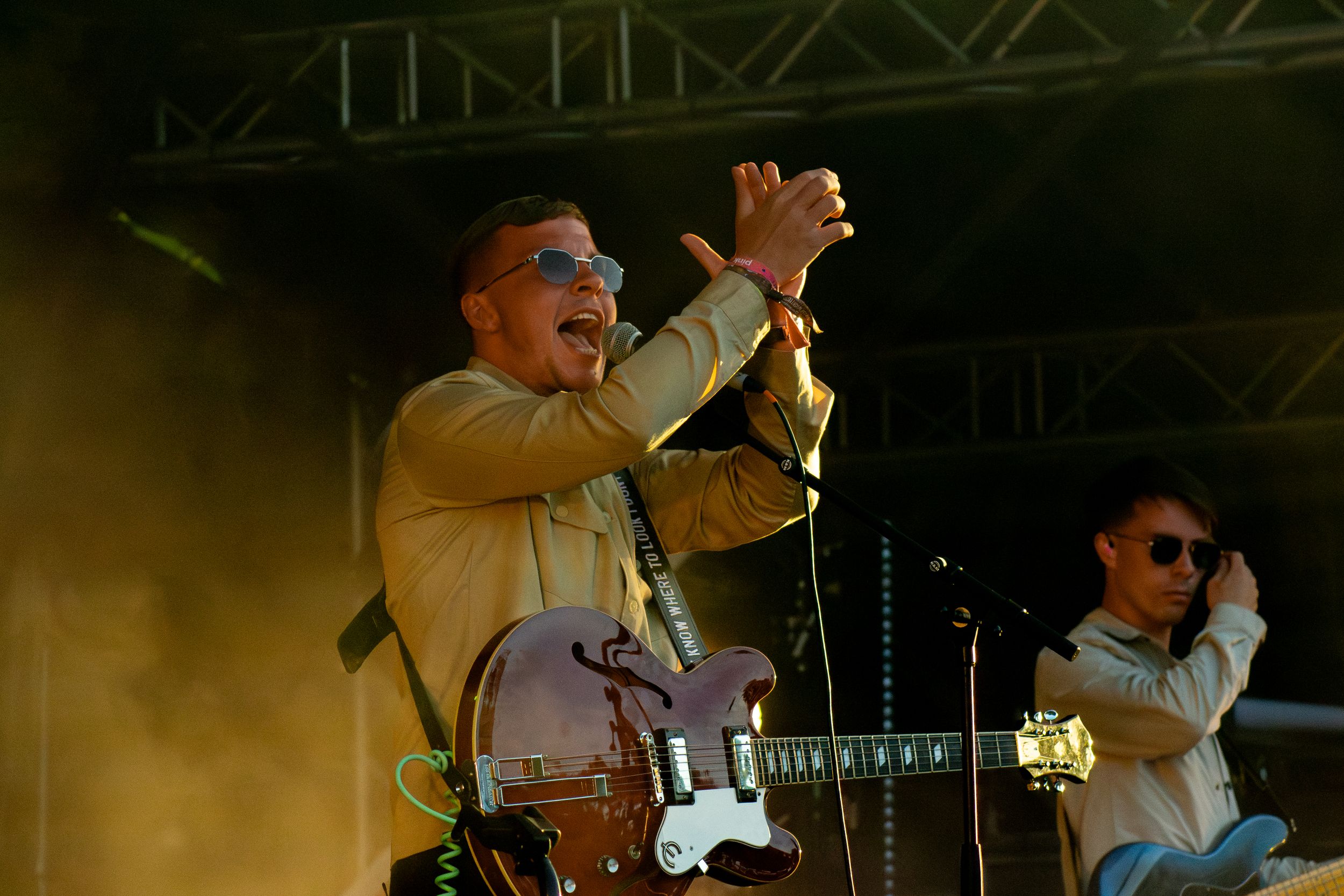 Musician plays electric guitar on stage bathed in warm golden lighting at outdoor concert performance.