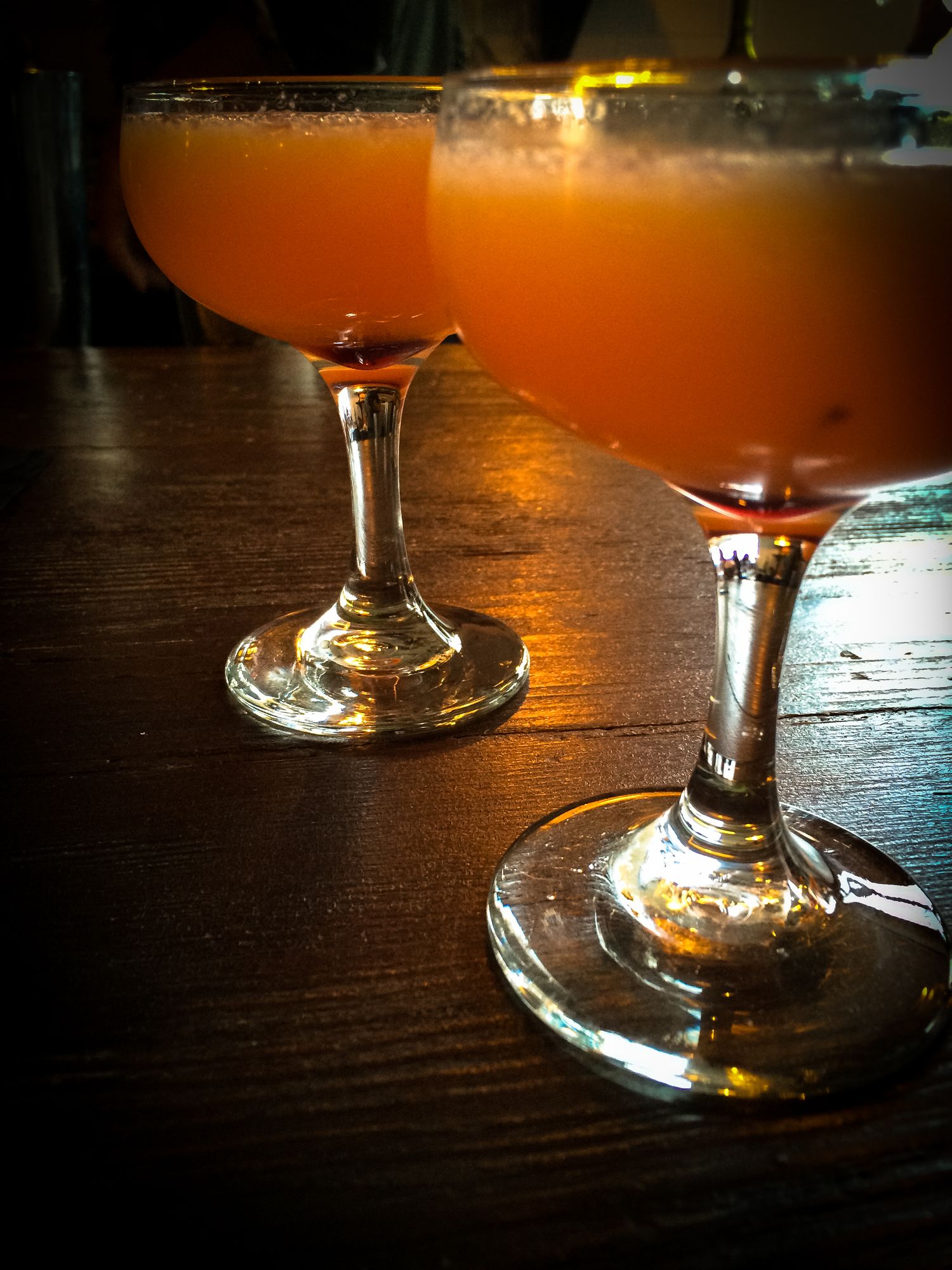 Two orange cocktails in stemmed glasses on dark wooden bar counter.