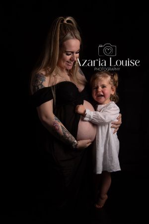 A touching maternity portrait shows a mother in a black dress and her excited young child against a dark background.
