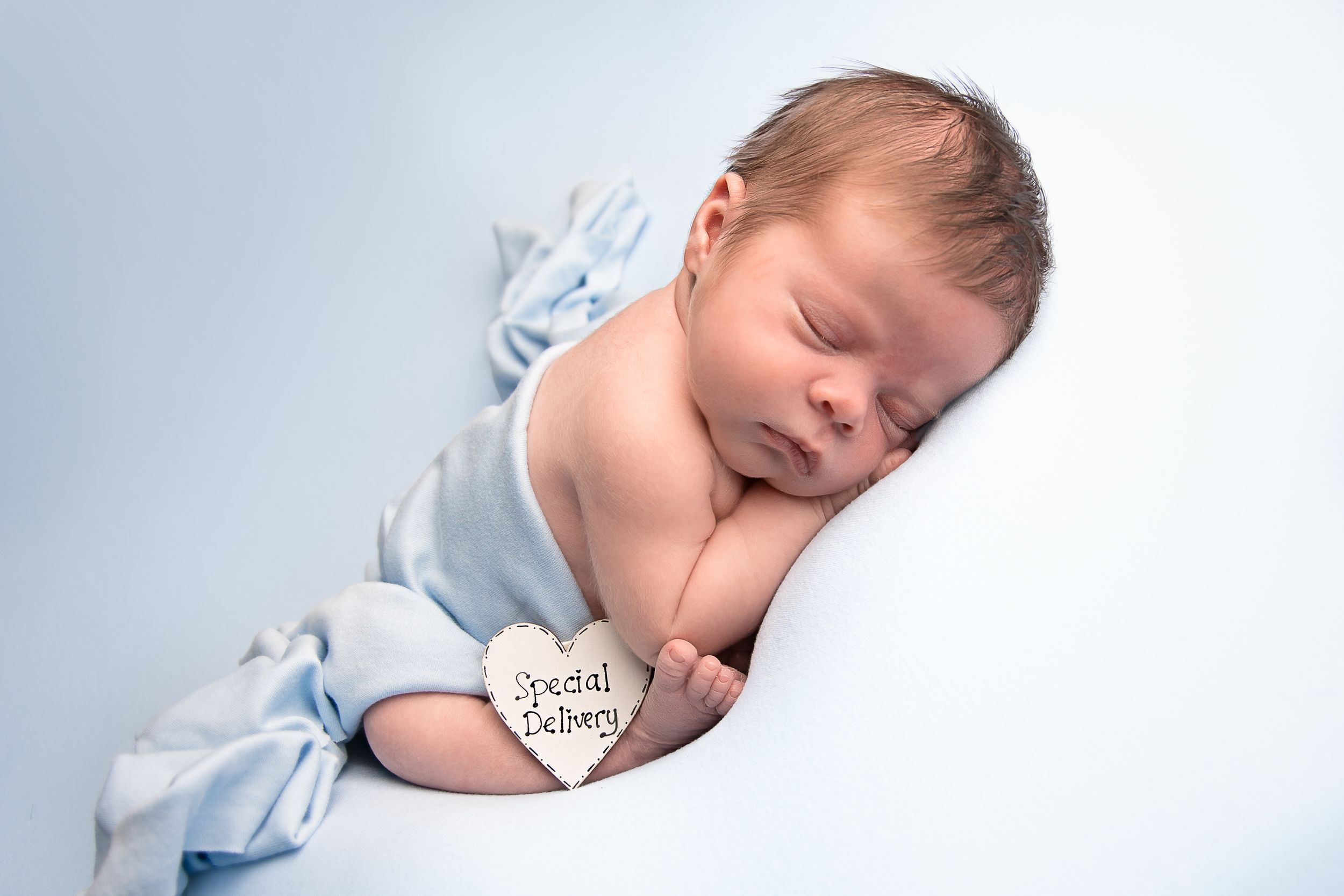 A newborn baby sleeps peacefully wrapped in a light blue blanket while resting on a white background.