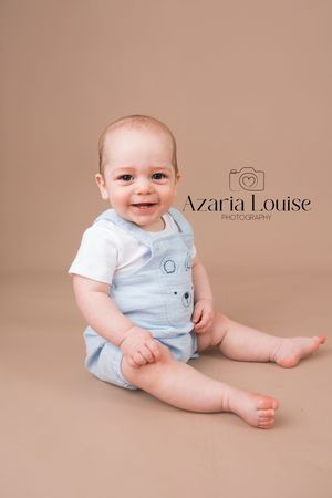 A baby wearing a light blue and white outfit sits on a beige backdrop in a professional studio portrait.