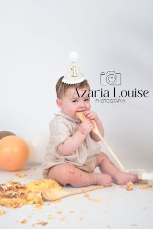 Baby in beige outfit and birthday hat sits on floor eating cake during first birthday photo shoot with orange balloon.