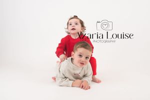 Two young children in studio photography pose, with one in red and one in beige laying on white backdrop.