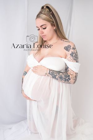 A maternity photo showing someone in a white off-shoulder dress with ornate sleeve tattoos against a light background.
