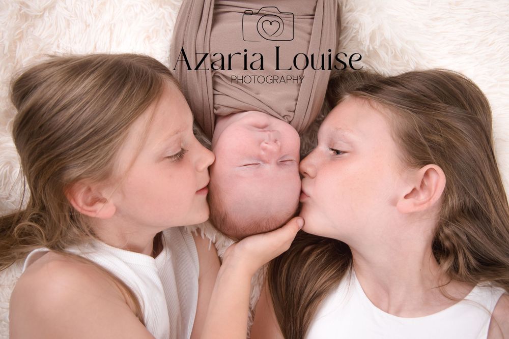 Two children embracing and kissing a baby from both sides while lying down together on a soft beige surface.